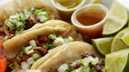 Mexican tacos in a container with limes and tomatoes.