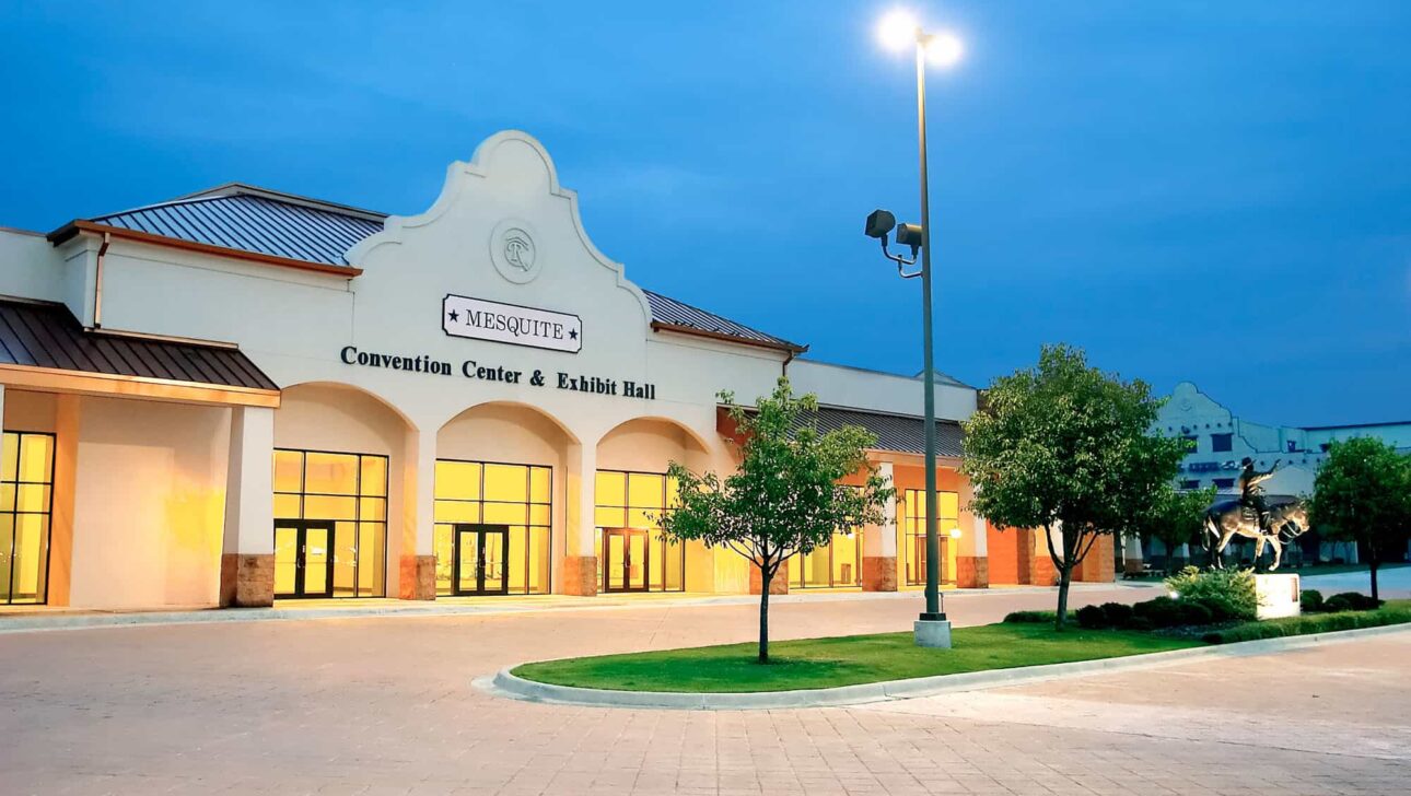 A well-lit entrance to the mesquite convention center & exhibit hall at twilight with a clear sky and a paved open space lined with young trees.