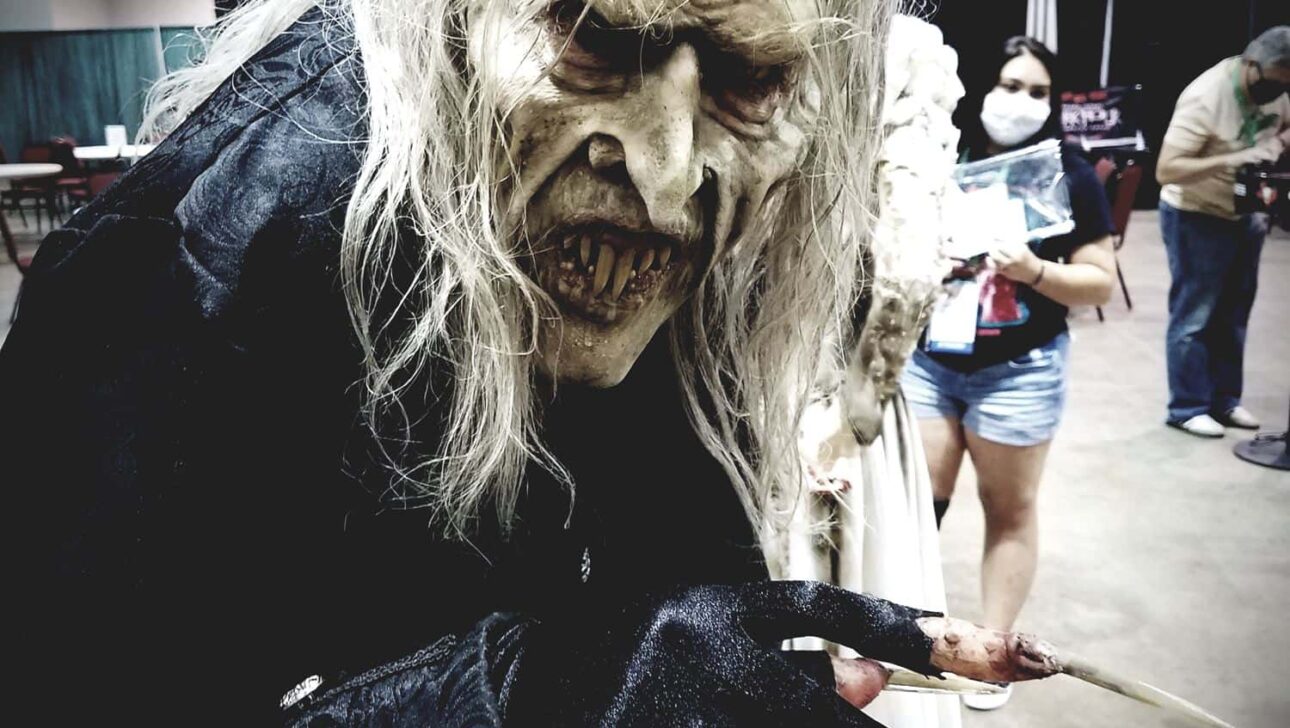 Person wearing a horror-themed costume with a grotesque mask and clawed gloves at a convention.