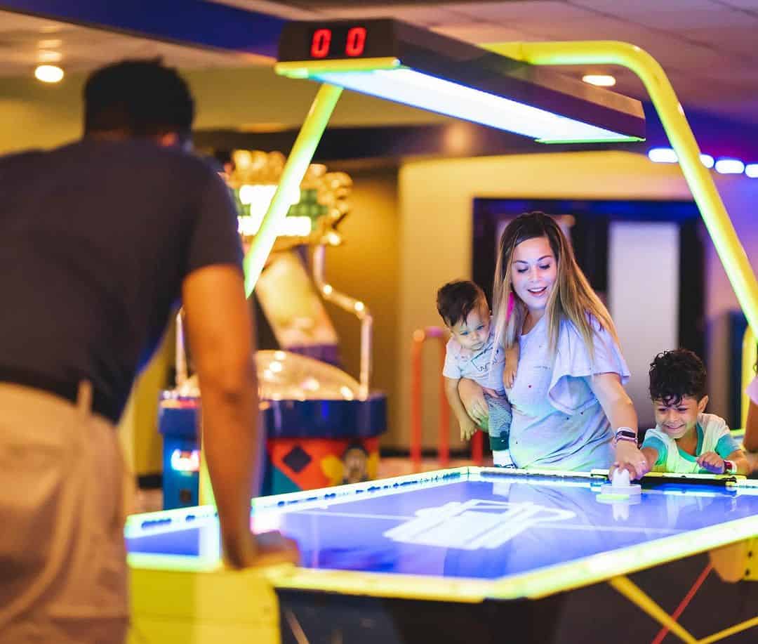 A woman and two young children are playing air hockey against a male opponent in an arcade setting.