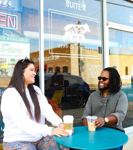 Two individuals are seated at a small green table outside a cafe, enjoying coffee and a conversation.