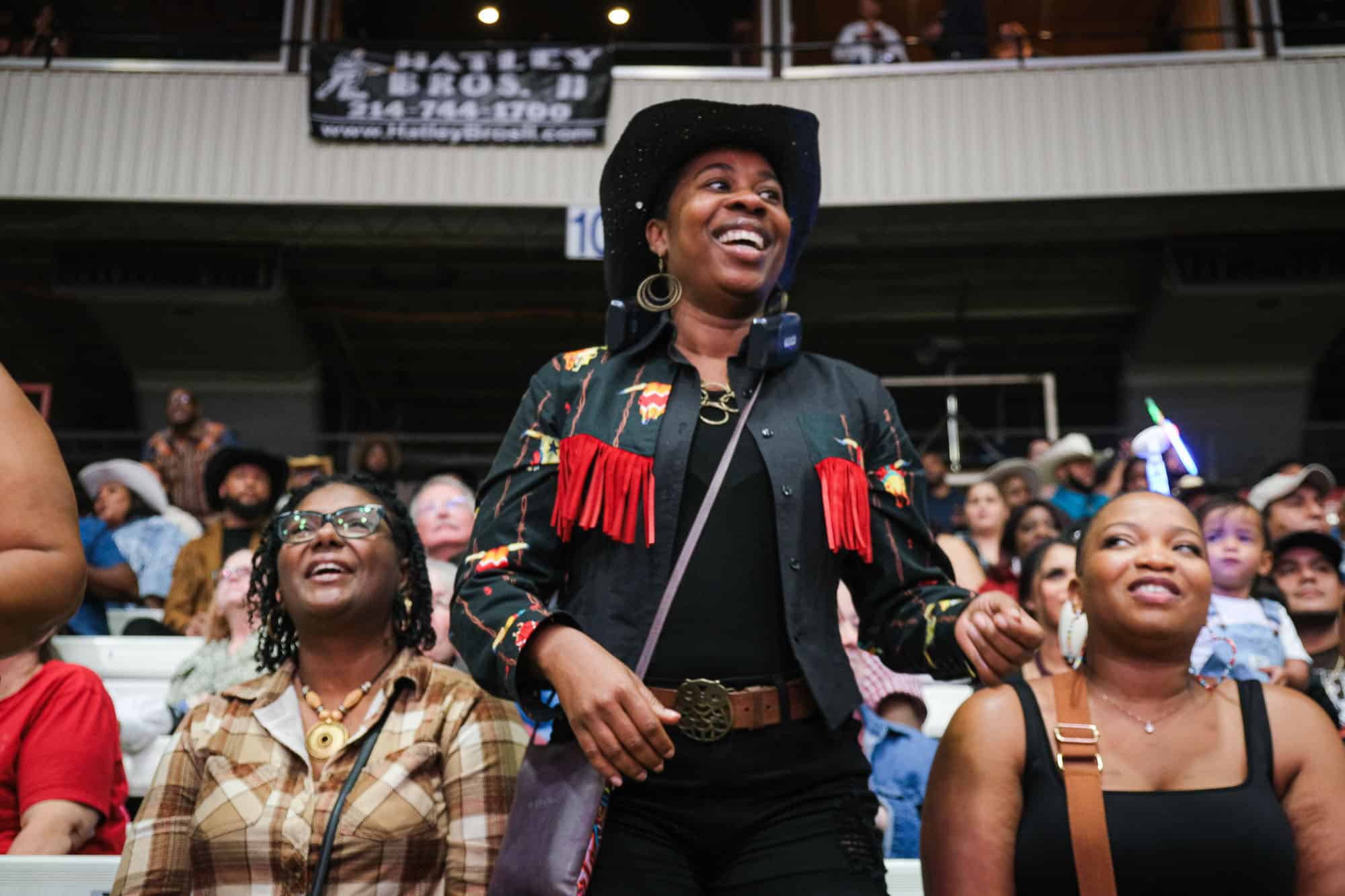 A woman in a black hat and embroidered jacket standing and smiling at an event, with other attendees seated around her.