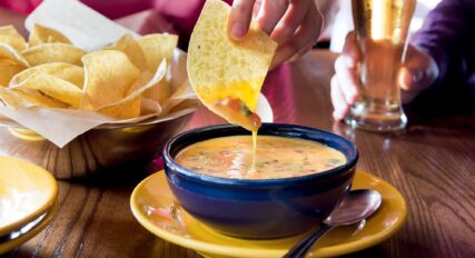 A person is dipping a tortilla chip into a bowl of cheese dip at a wooden table, with a glass of beer and additional chips in the background.
