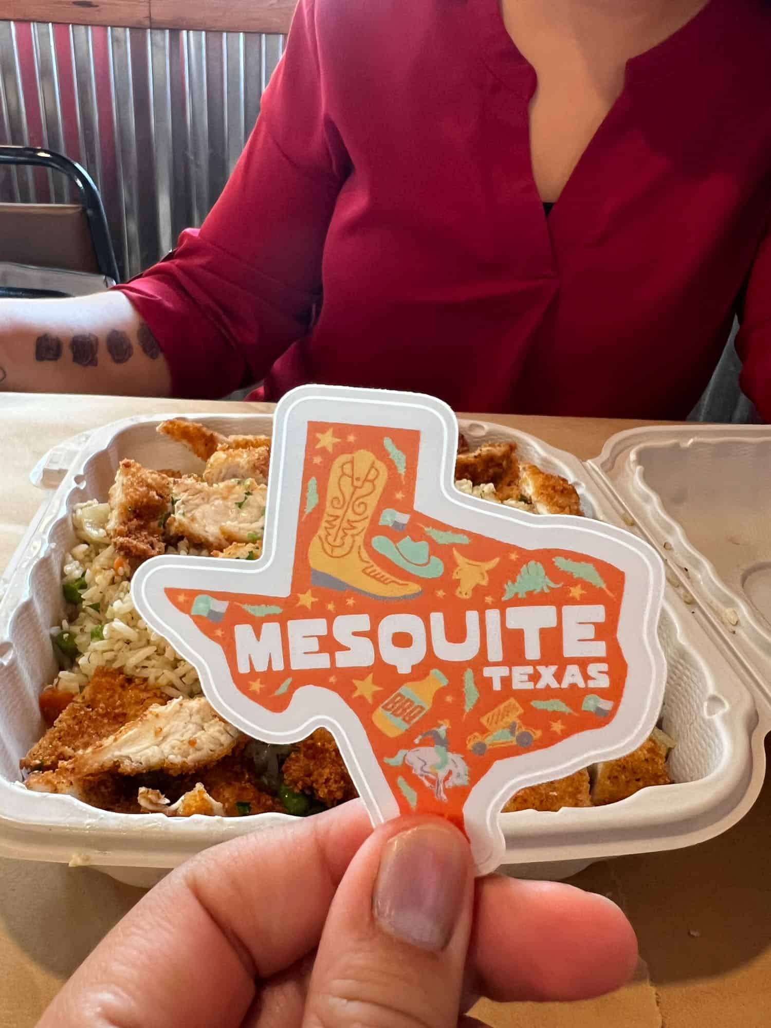 A person is holding a sticker with "mesquite texas" printed on it in front of a takeout container of food, with another person visible in the background.