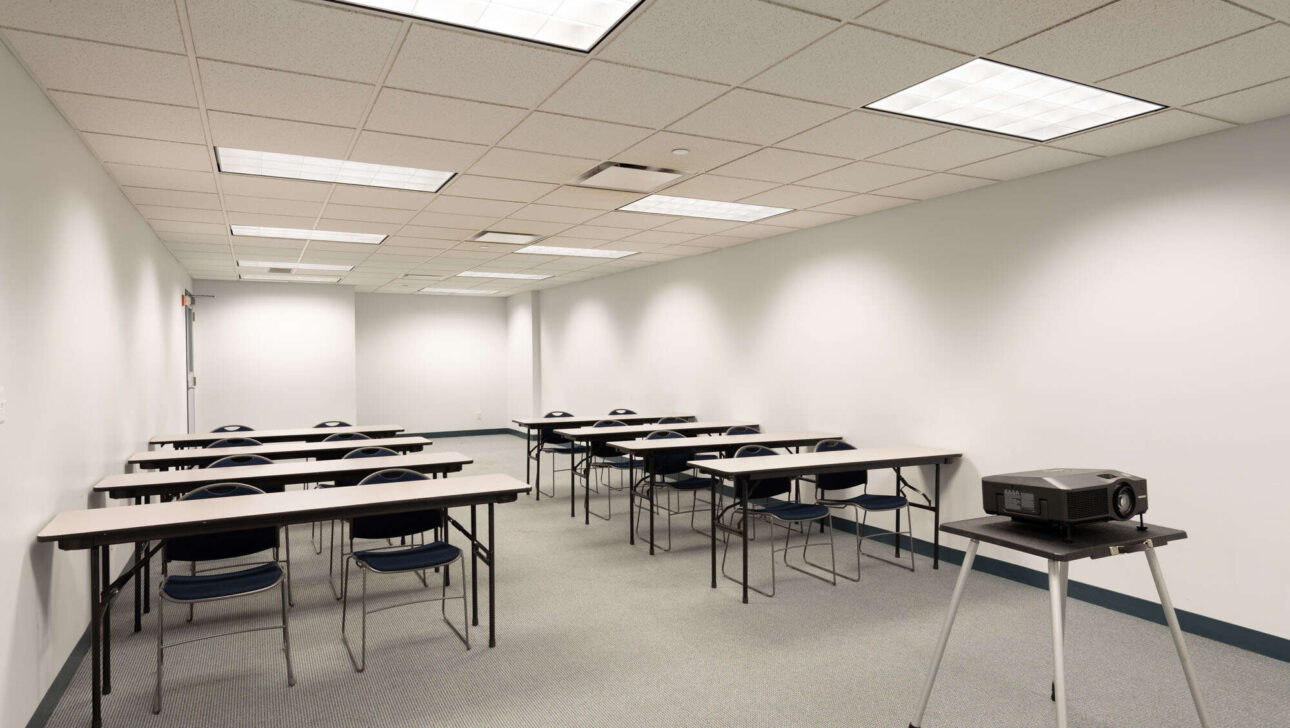 An empty classroom with rows of desks and chairs, a projector on a stand, and a whiteboard at the front, illuminated by fluorescent lighting.