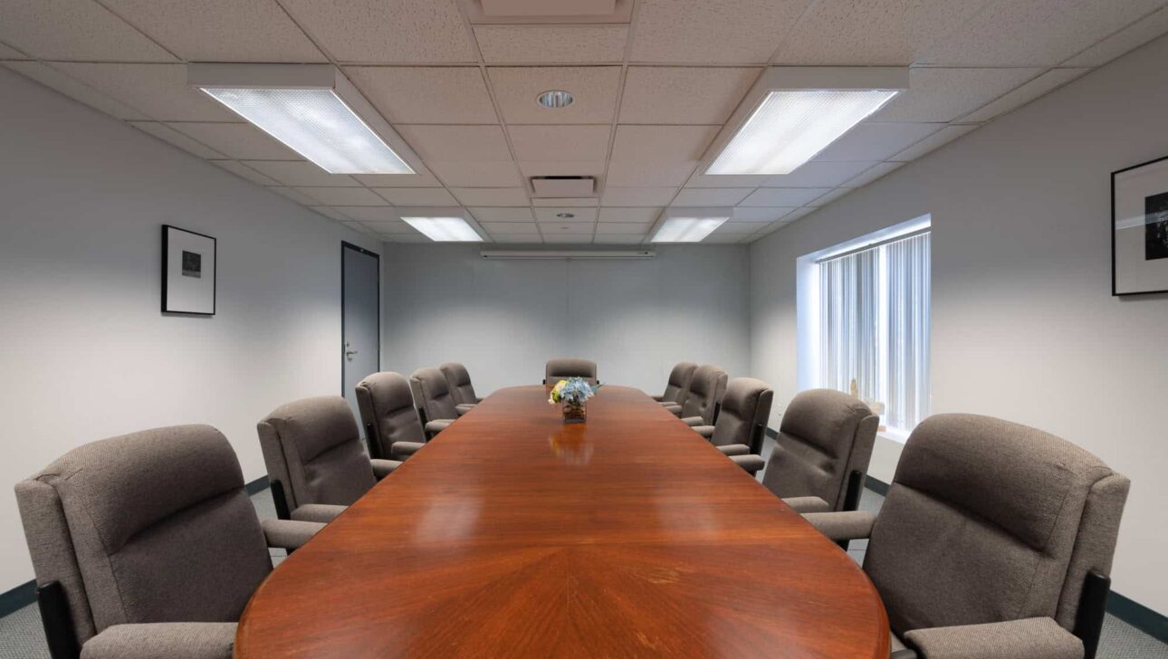 A conference room with a long wooden table surrounded by grey upholstered chairs, fluorescent lighting on the ceiling, and framed pictures on the walls.