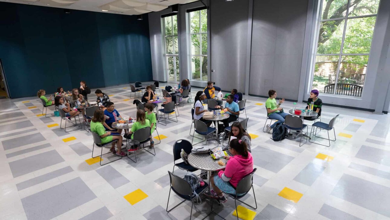 Groups of children are seated at round tables participating in an indoor activity or class in a spacious room with large windows and a checkered floor.