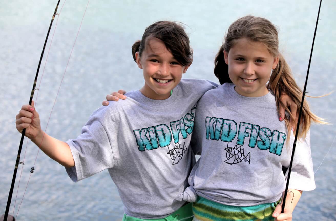 Two girls wearing "KID FISH" shirts stand side by side, smiling and holding fishing rods by a body of water.