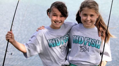 Two girls wearing "KID FISH" shirts stand side by side, smiling and holding fishing rods by a body of water.