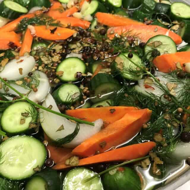 Sliced cucumbers, carrots, onions, dill, and spices soaking in a clear pickling brine inside a large round container.