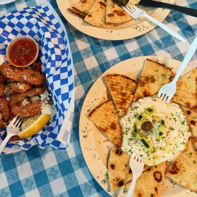 Plates of pita bread with hummus and a basket of fried zucchini sticks with marinara sauce on a blue checkered tablecloth.
