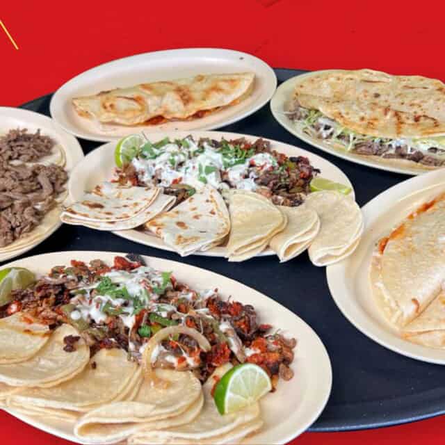 A variety of Mexican dishes, including quesadillas, tacos, grilled meat, and garnishes, arranged on round plates over a black tray with a red background.