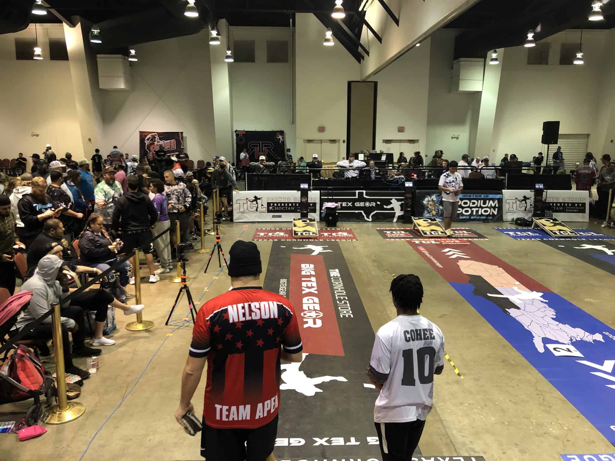 A large indoor competition event features two contestants, Nelson and Correa, preparing to participate. Spectators line the area, and the venue is equipped with banners and various setups for the event.
