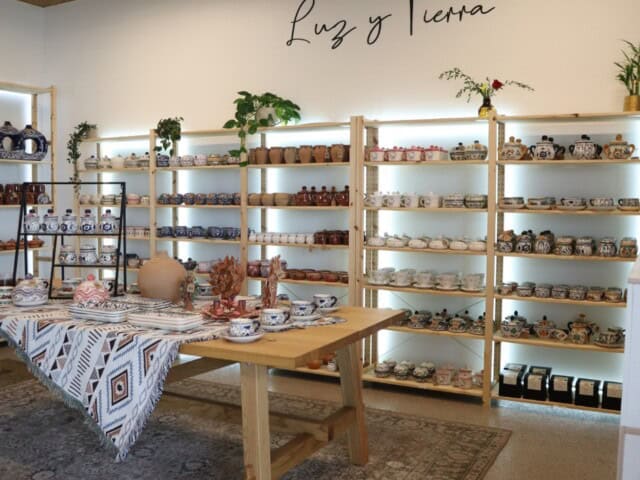 A pottery store featuring shelves of various ceramic items, including bowls and mugs. A wooden table at the center holds additional pottery pieces and is adorned with a patterned cloth.