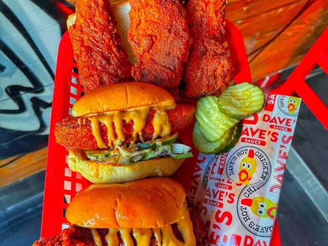 A red basket contains two spicy chicken sandwiches topped with sauce and coleslaw, accompanied by three fried chicken tenders and pickle slices.