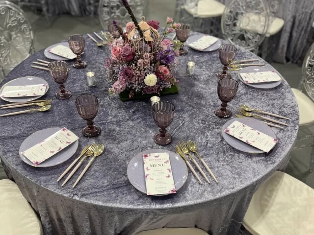 Elegant table setting with purple and pink floral centerpiece, transparent chairs, purple glassware, gold cutlery, and menu cards on each plate.