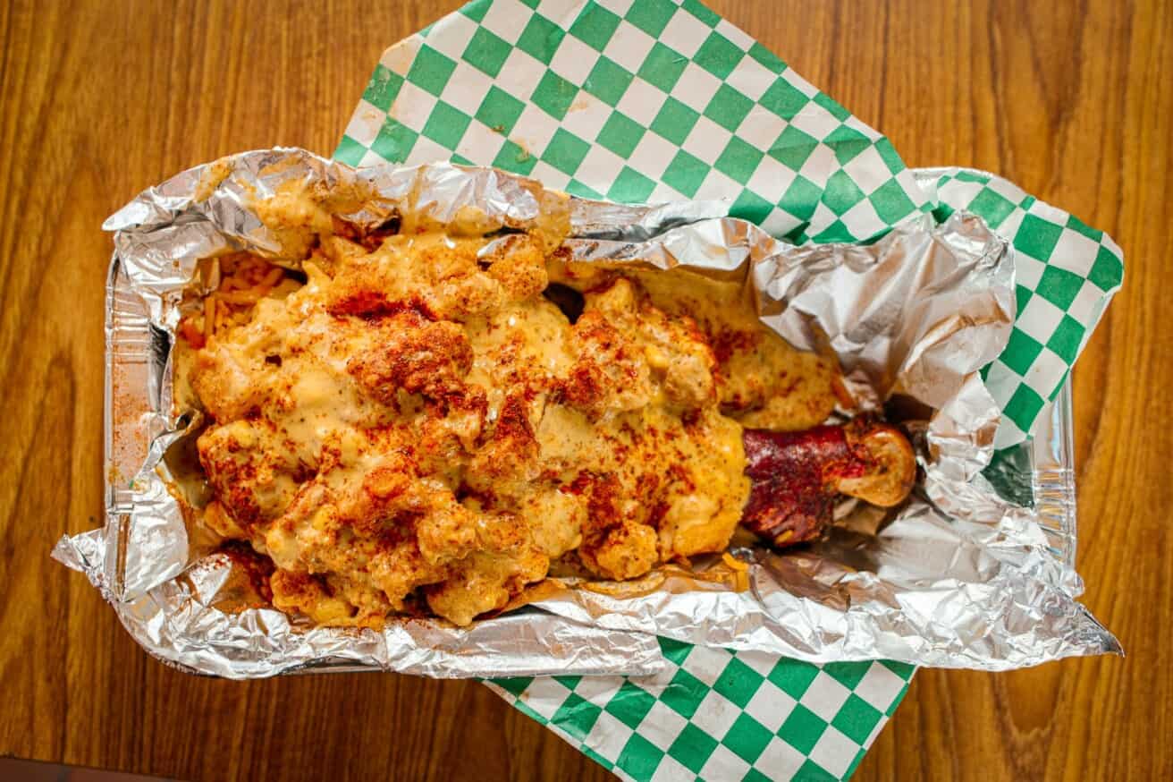 Barbecue ribs topped with melted cheese and seasoning, served on a foil tray with a green checkered paper liner, set on a wooden table.