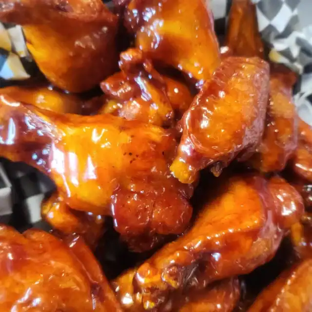 Close-up of a pile of glazed chicken wings coated in a shiny, rich sauce, served on black and white checkered paper.