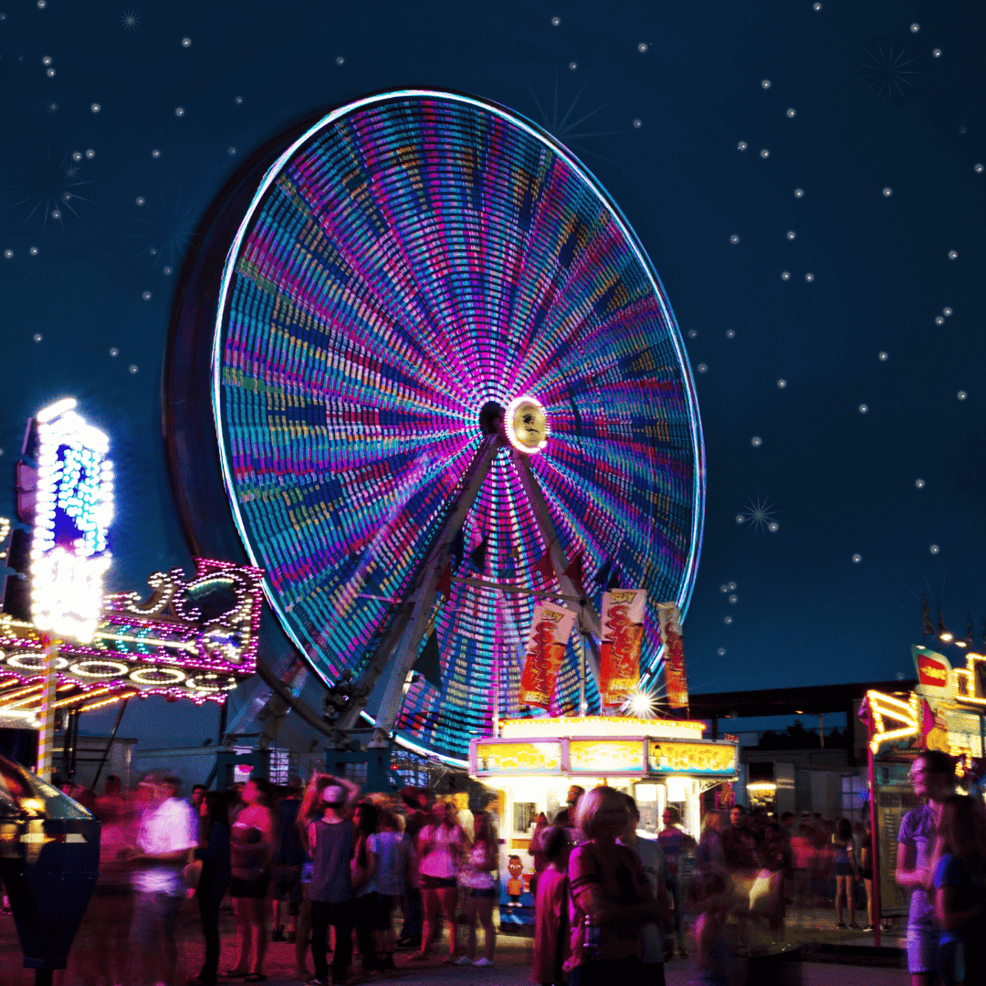 A brightly lit Ferris wheel spins at night, surrounded by blurred crowds and illuminated stalls. The sky is dark with scattered small stars.