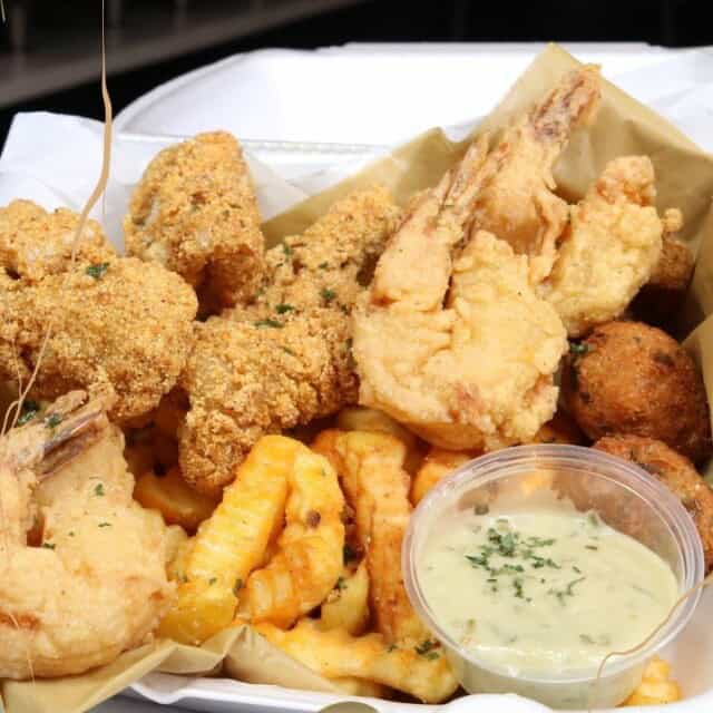 A takeout container filled with fried shrimp, fish, crinkle-cut fries, and hush puppies, accompanied by a cup of creamy dipping sauce.
