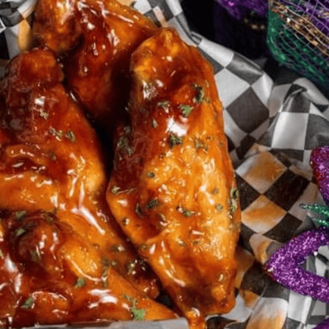 Chicken wings coated in a glossy barbecue sauce are served on black and white checkered paper, with Mardi Gras beads visible in the background.