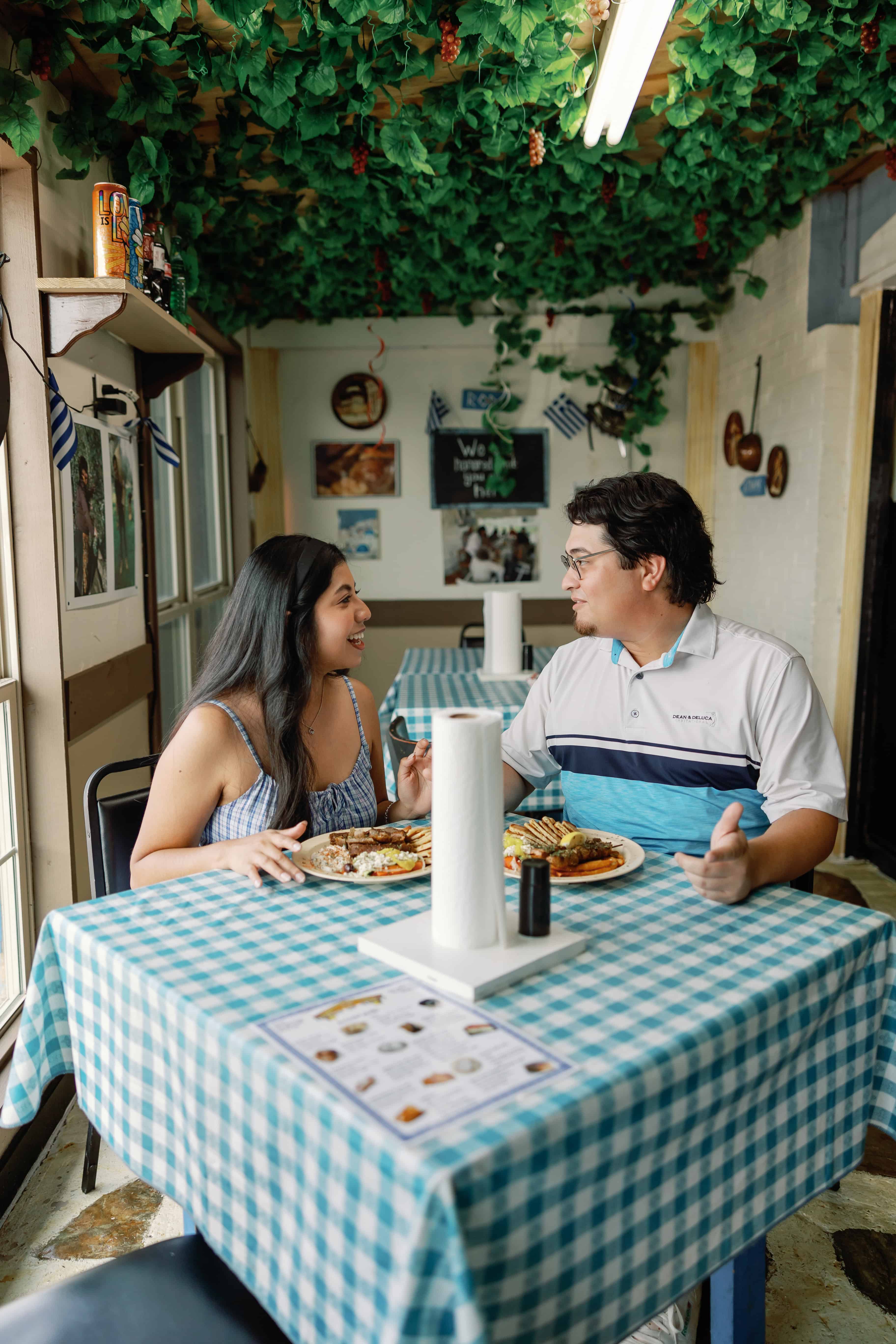 Two people sitting at a checkered table in a restaurant with green foliage on the ceiling, engaging in conversation while dining. Menus and Greek flags are visible around the room.