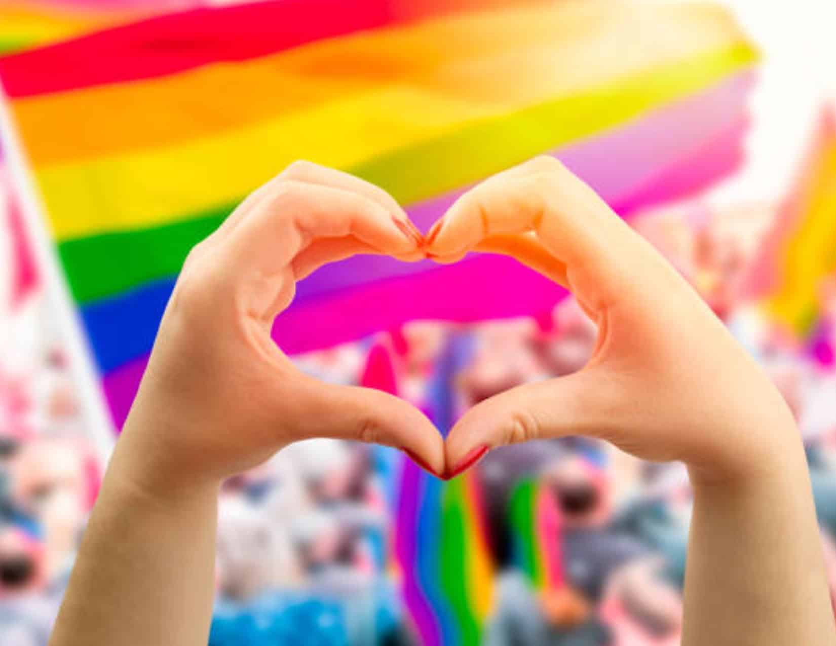 Hands forming a heart shape in front of a blurred crowd with a large rainbow flag in the background.