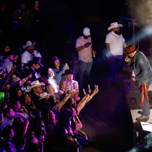 Performer singing on stage in front of a cheering crowd; audience members are taking photos, some wearing cowboy hats.