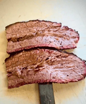 Two thick slices of cooked brisket with a visible smoke ring and a knife underneath, displayed on a light-colored cutting board.