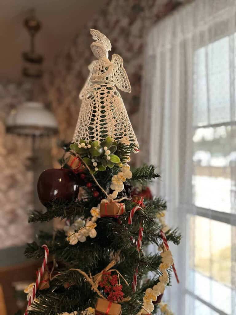 A Christmas tree topped with a crocheted angel, decorated with red ornaments, wrapped gifts, candy canes, and garland, next to a window with sheer curtains.