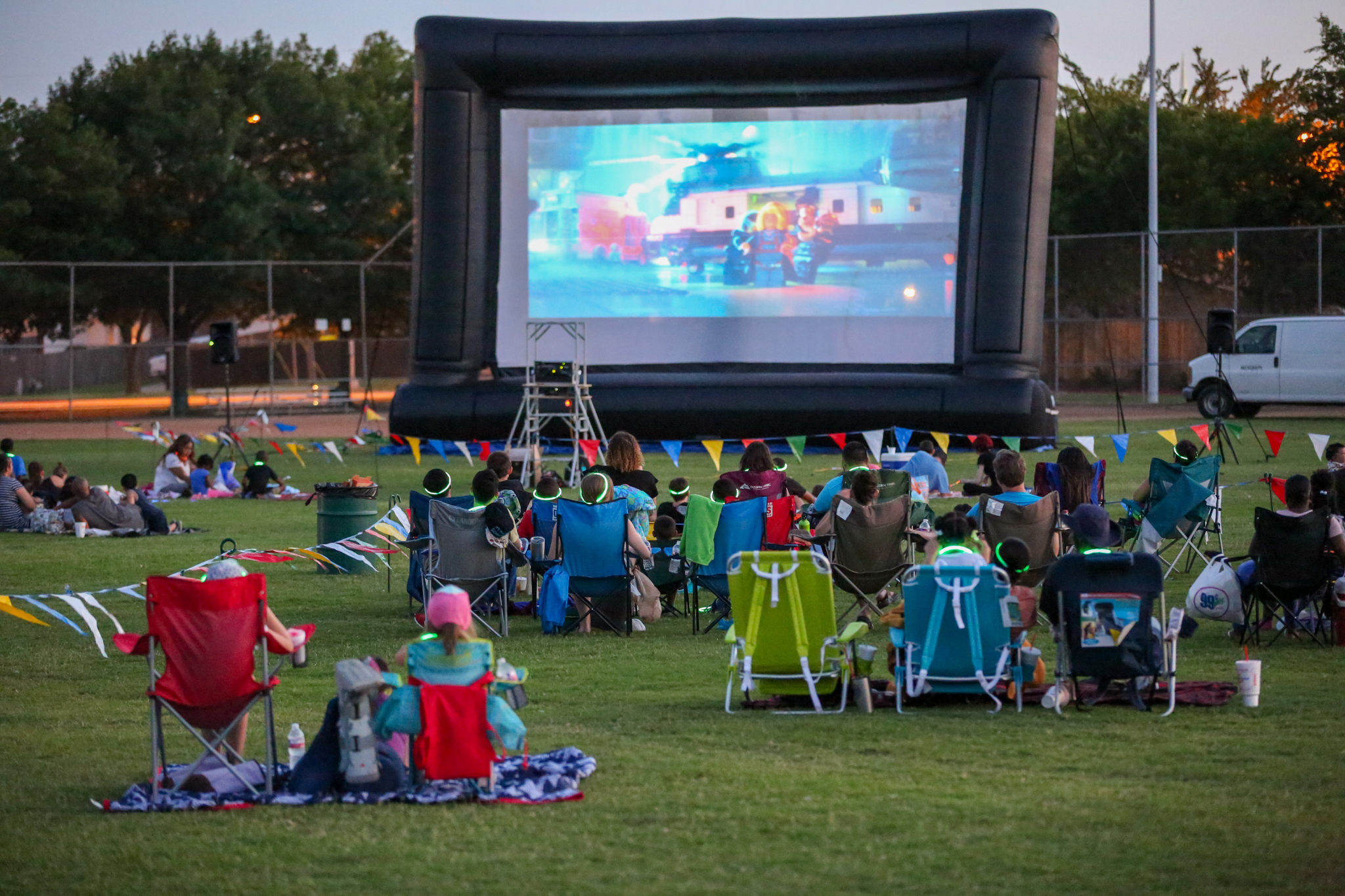 People sit on lawn chairs and blankets watching a movie projected on a large outdoor screen in a park during the evening.