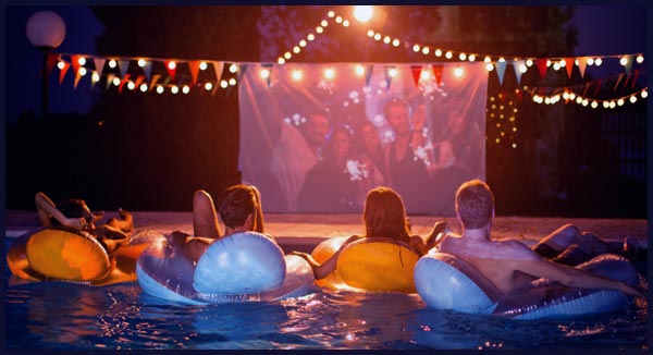 Four people sit on pool floats watching a movie projected on a screen at night, with string lights and bunting decorations overhead.