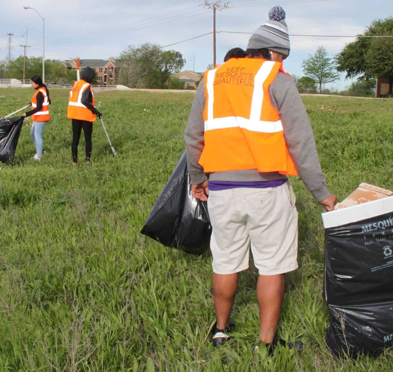 Four people wearing orange safety vests pick up trash and debris in a grassy field as part of an outdoor cleanup effort.