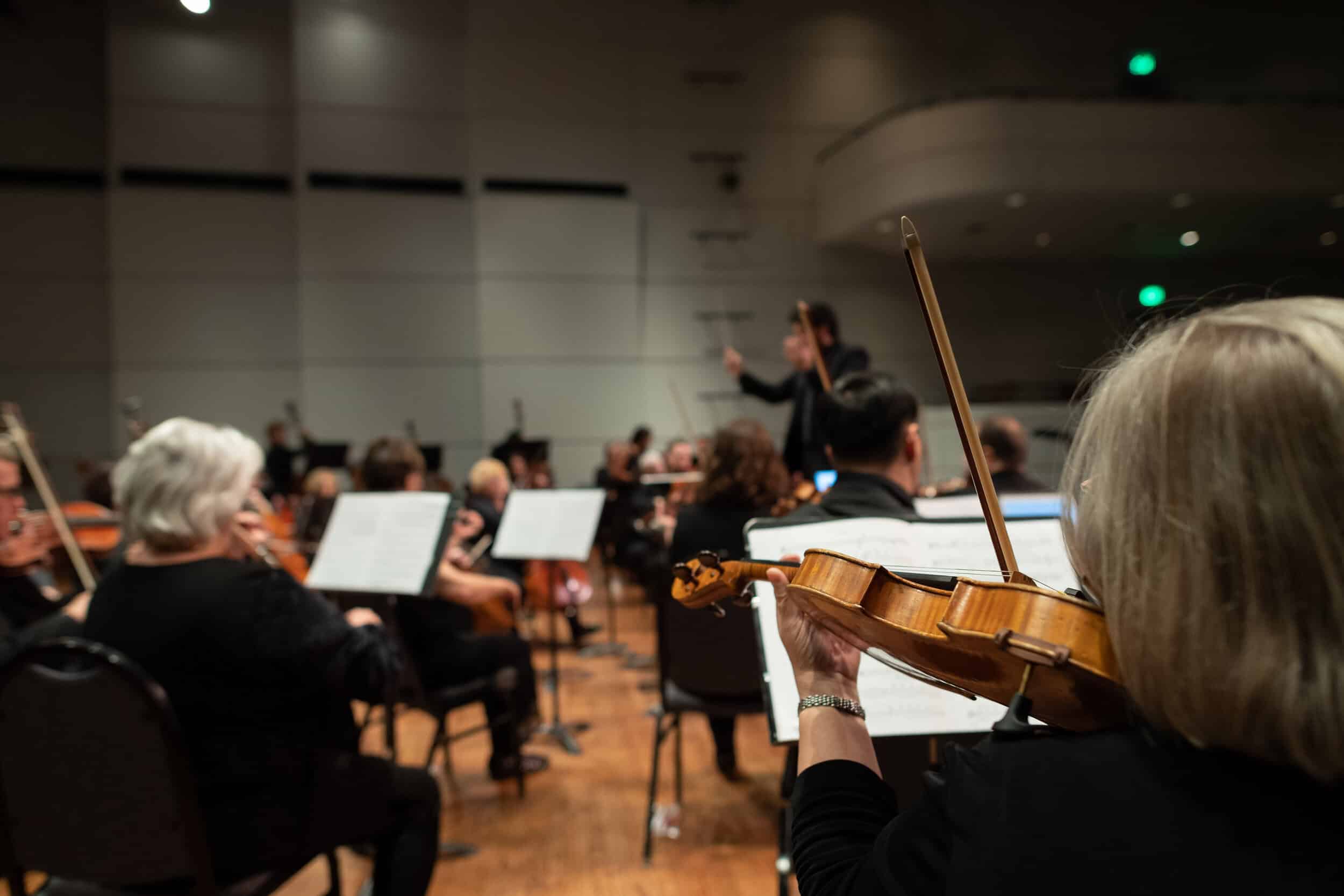 Orchestra musicians seated on stage, playing string instruments, with a conductor leading in the background.