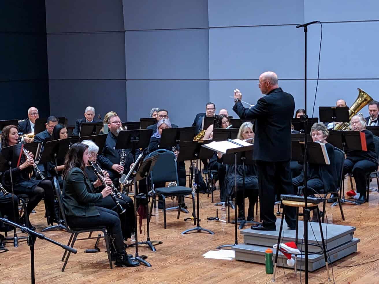 A conductor leads a seated orchestra of musicians playing various instruments on a stage with music stands and microphones.
