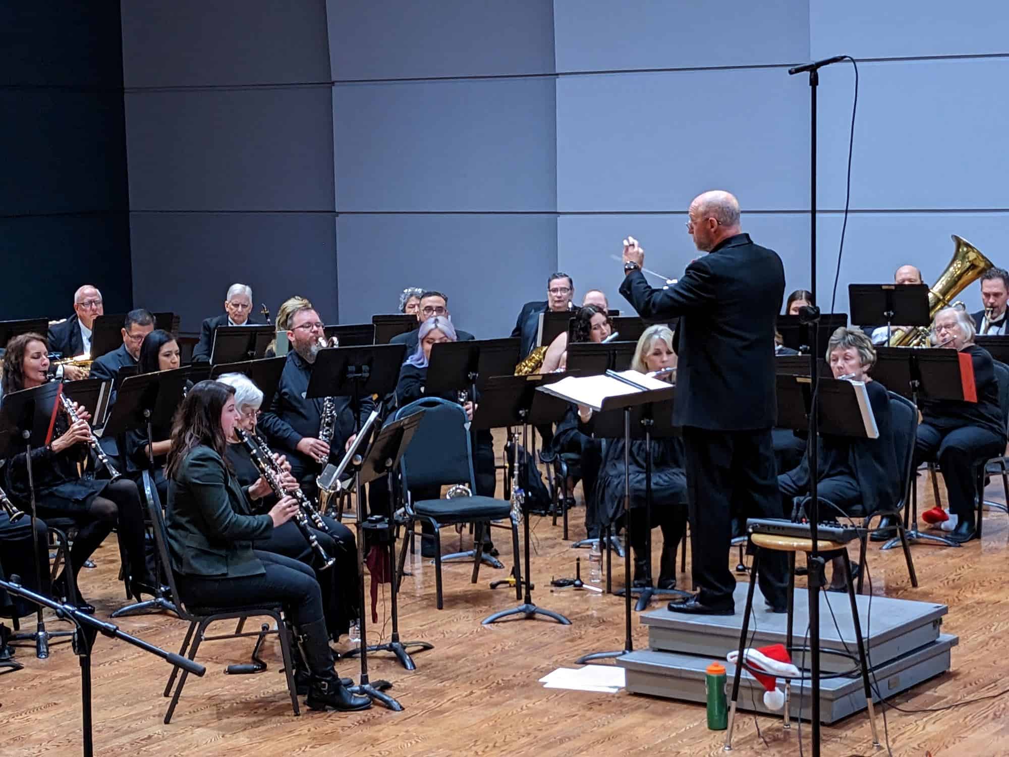A conductor leads a seated orchestra of musicians playing various instruments on a stage with music stands and microphones.