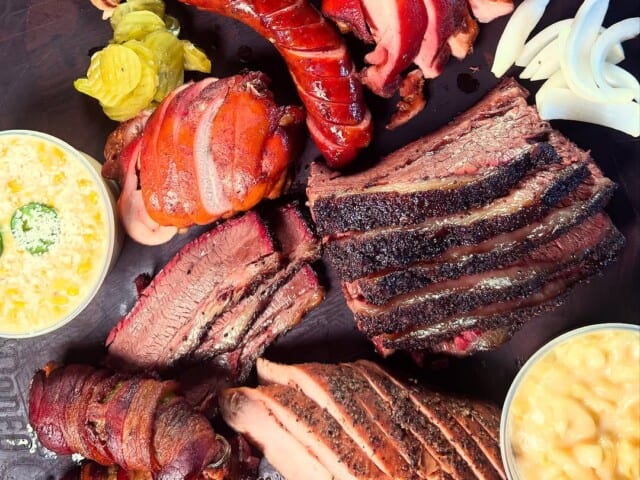 An assortment of barbecued meats including brisket, sausage, and bacon-wrapped items, served with pickles, onions, macaroni and cheese, beans, and corn.
