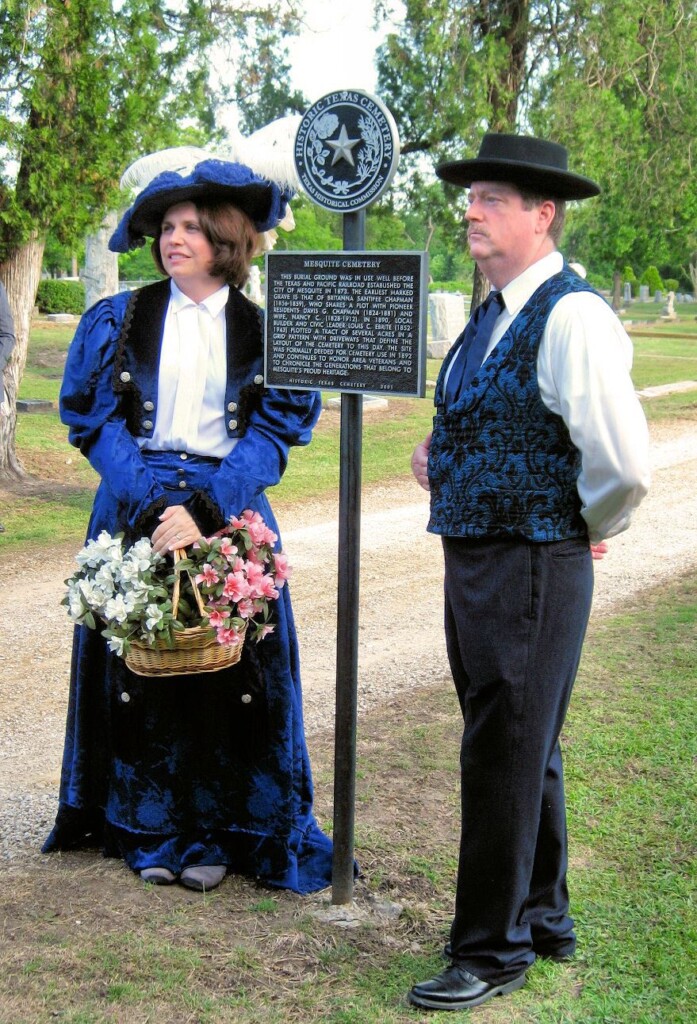 Two adults in period costumes stand by a historical marker in a cemetery; one holds a basket of flowers. Trees and headstones are visible in the background.