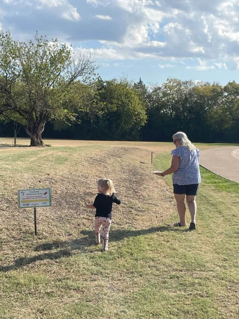 An older woman and a young girl walk on a grassy area near a sign, with trees and a partly cloudy sky in the background.