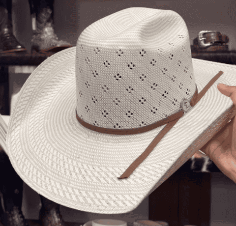 A cream-colored straw cowboy hat with a patterned brim and crown, brown hatband, and decorative perforations, held in a hand in front of a display of boots and belts.