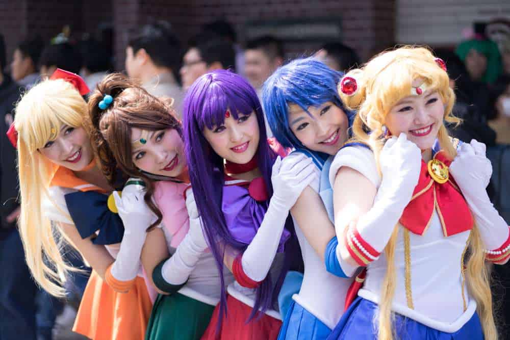 Five people dressed in colorful Sailor Moon character costumes pose together outdoors, smiling at the camera with a crowd in the background.