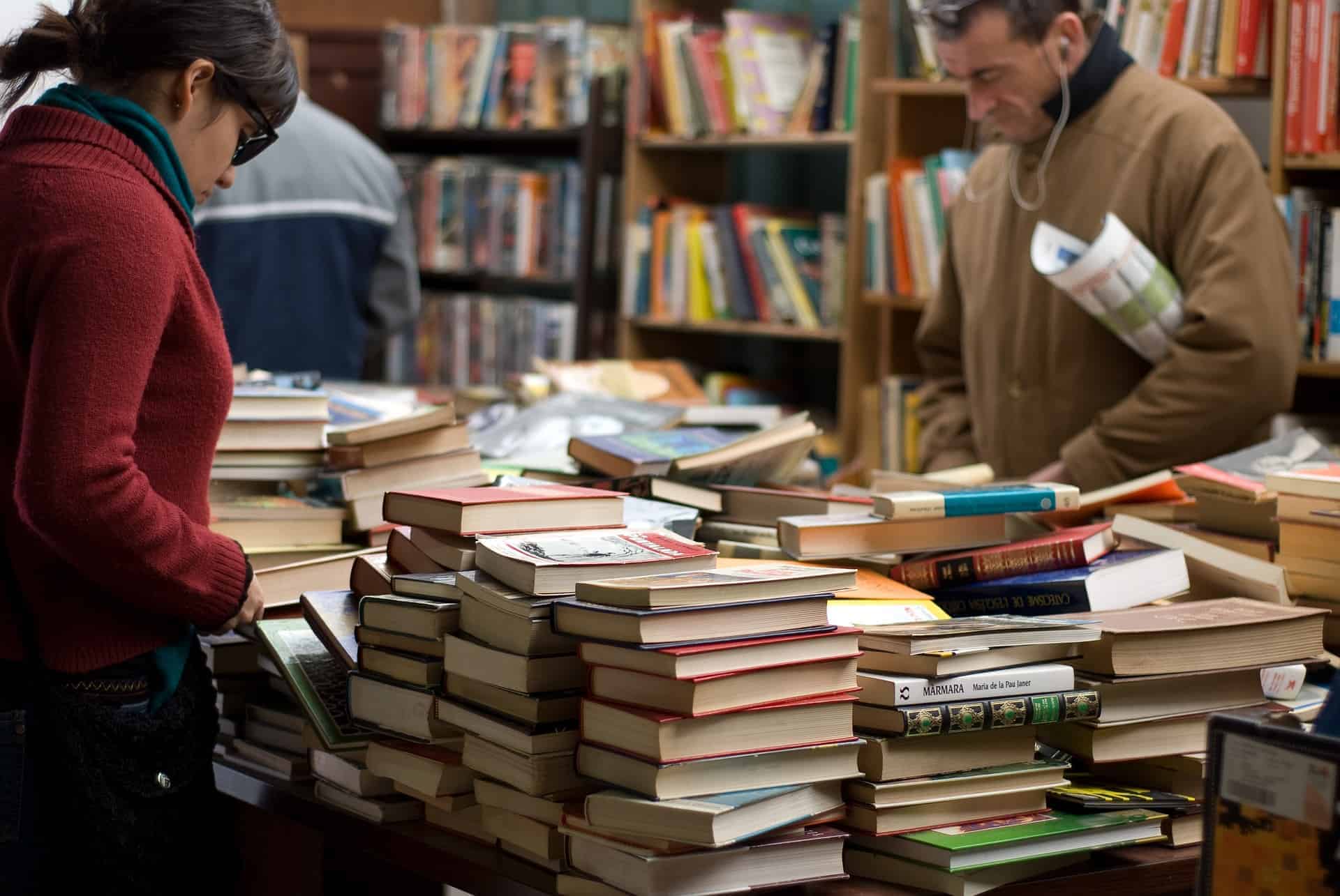 Two people browse through stacks of books on tables inside a bookstore or library, with shelves of more books in the background.
