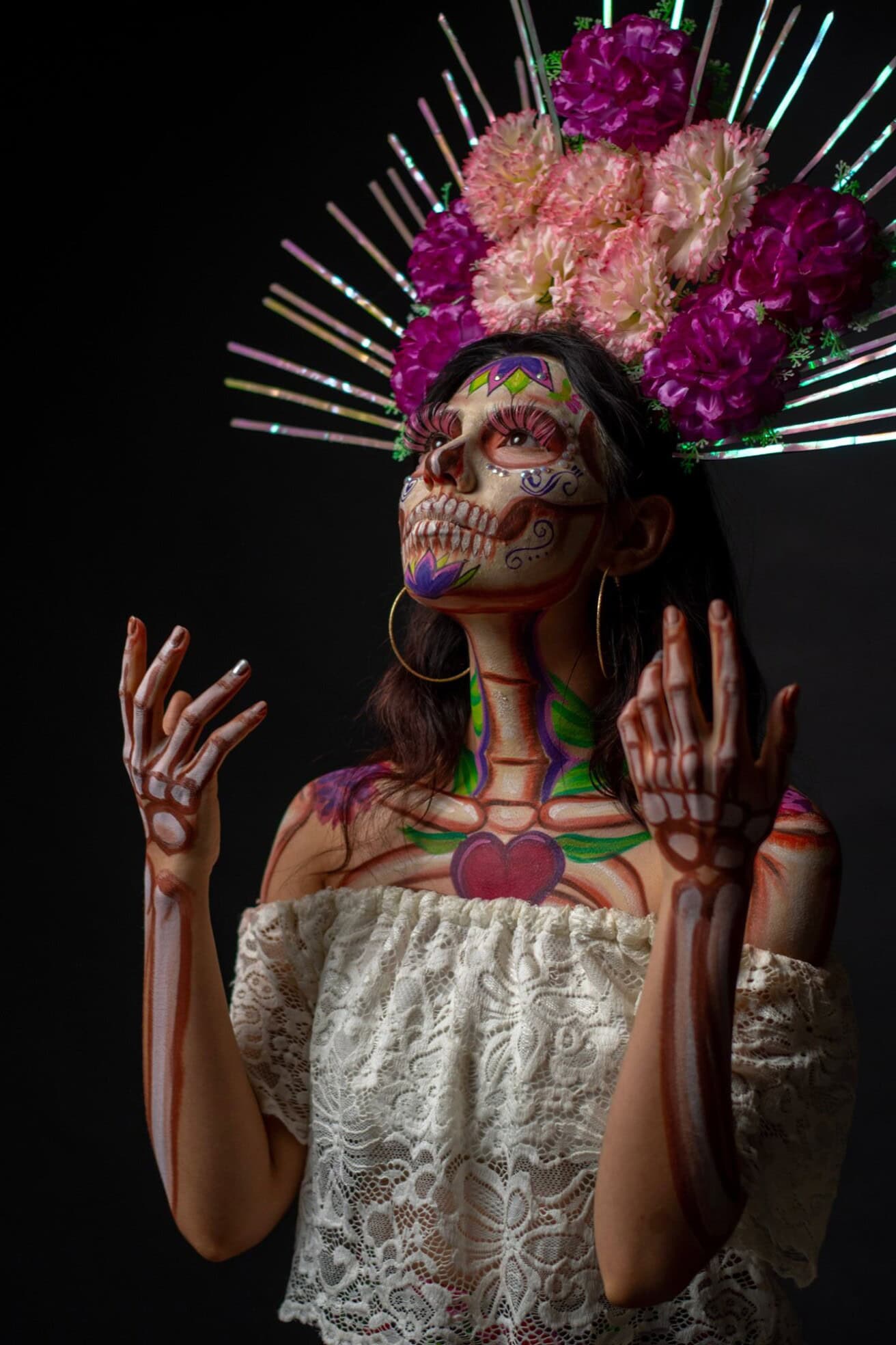 A person wearing a white lace top poses with Day of the Dead skeleton makeup, colorful flowers, and decorative metallic spikes on their head against a dark background.