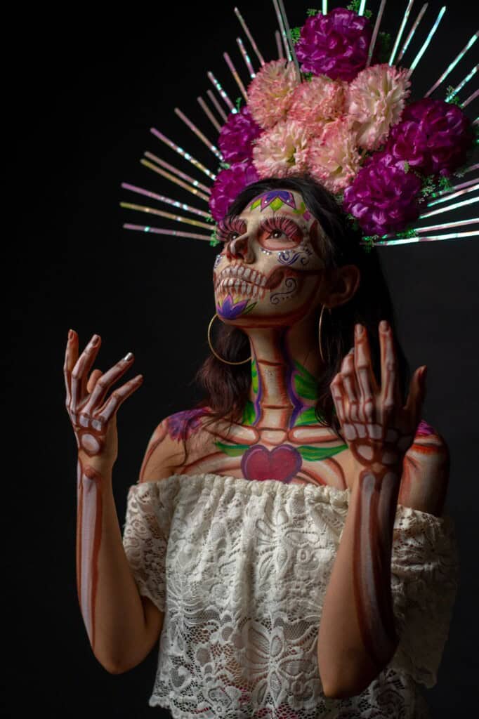 A person wearing a white lace top poses with Day of the Dead skeleton makeup, colorful flowers, and decorative metallic spikes on their head against a dark background.