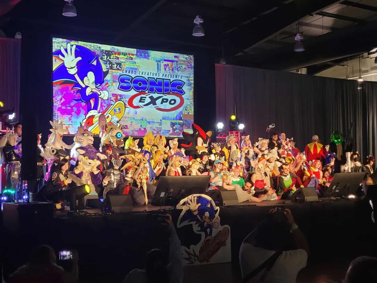 A large group of people in Sonic the Hedgehog costumes pose on stage at Sonic Expo, with a colorful Sonic Expo sign displayed in the background.