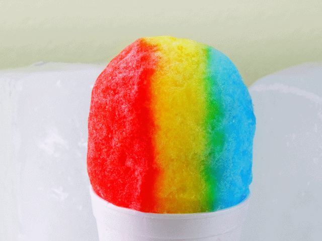 A snow cone in a white foam cup with red, yellow, green, and blue syrup, placed in front of a background with large ice blocks.