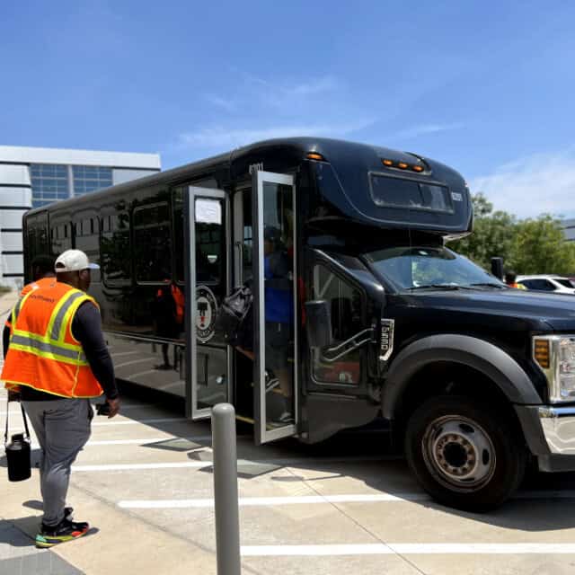 A black shuttle bus with its door open waits at a curb while people in safety vests walk nearby on a sunny day.