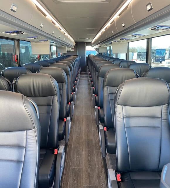 Interior of a modern coach bus with rows of empty black leather seats, wood-style flooring, and overhead storage compartments.