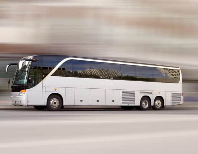 A white coach bus drives quickly along a city street, with the background blurred to indicate motion.