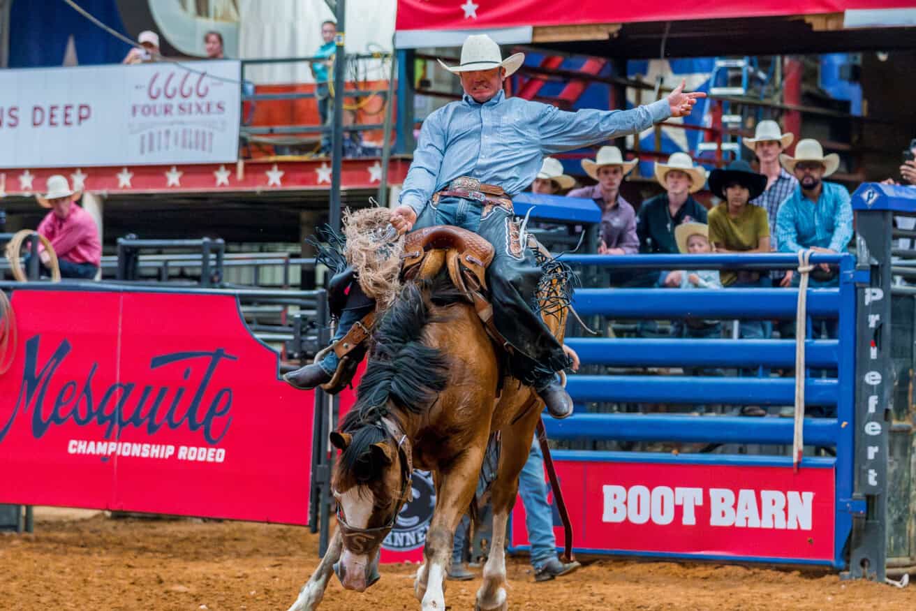 A rodeo rider wearing a cowboy hat and blue shirt rides a bucking horse in an arena, with a cheering crowd and sponsor signs in the background.